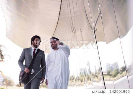 Two businessmen walking and talking by a modern glass building, one in a suit and one in a white robe Two businessmen walking and talking by a modern glass building, one in a suit and one in a white robe 142901996