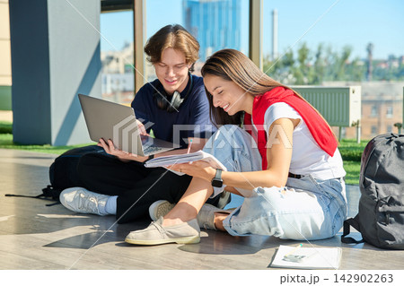 Two young students studying with laptop and notebook sitting on floor near big window 142902263