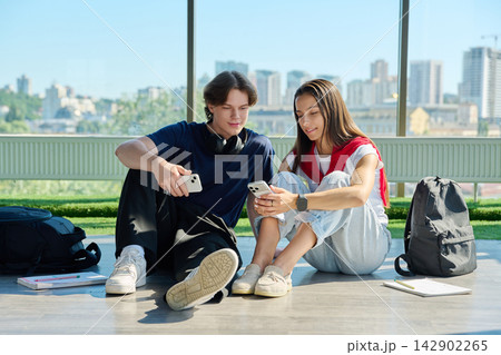 Two young students sitting on floor and looking at smartphones near window 142902265