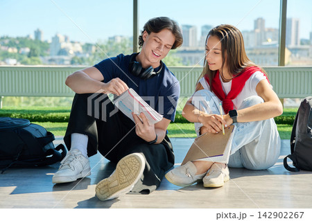 Two young students studying together with notebooks sitting on floor near big window Two young students studying together with notebooks sitting on floor near big window 142902267
