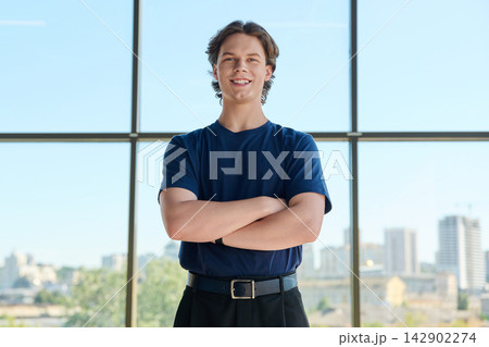 Smiling young man standing with crossed arms near large window with city view 142902274