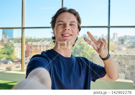 Happy young man taking selfie and showing victory sign near large window Happy young man taking selfie and showing victory sign near large window 142902276