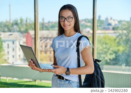 Smiling female student wearing eyeglasses holding laptop with city window background 142902280