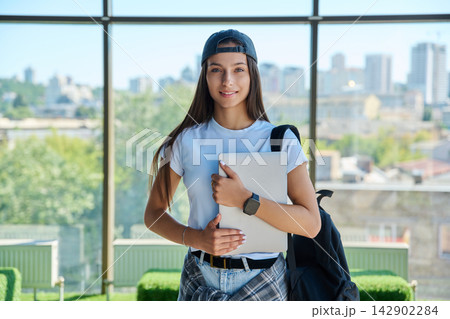 Smiling female student in cap holding laptop computer against city window Smiling female student in cap holding laptop computer against city window 142902284
