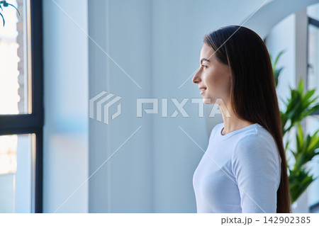 Profile portrait of serene young woman smiling near window indoors Profile portrait of serene young woman smiling near window indoors 142902385