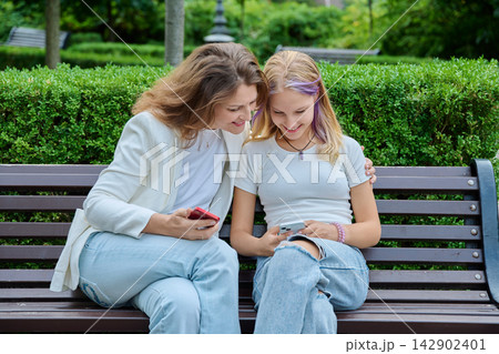 Mother leaning towards teenage daughter using smartphone on bench in park Mother leaning towards teenage daughter using smartphone on bench in park 142902401