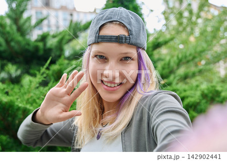 Smiling teenage girl with purple hair strands waving hand taking selfie outdoors Smiling teenage girl with purple hair strands waving hand taking selfie outdoors 142902441