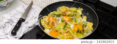 Fried zucchini is cooked with scrambled eggs in a pan on a stovetop. The scene shows a kitchen preparing a meal. Fresh ingredients are visible, creating a simple dish, banner 142902913