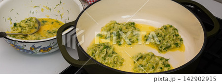Zucchini pancakes are being prepared in a pan on the stove. A bowl of pancake batter sits nearby. The kitchen has basic tools and a clean countertop, banner Zucchini pancakes are being prepared in a pan on the stove. A bowl of pancake batter sits nearby. The kitchen has basic tools and a clean countertop, banner 142902915
