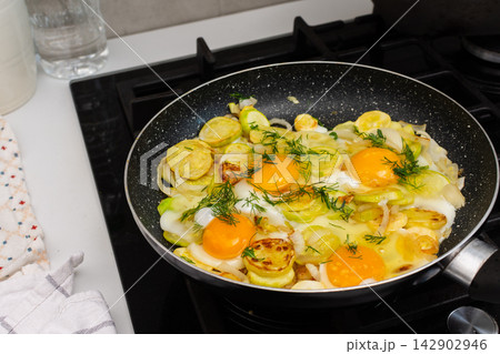 Fried zucchini is cooked with scrambled eggs in a pan on a stovetop. The scene shows a kitchen preparing a meal. Fresh ingredients are visible, creating a simple dish 142902946