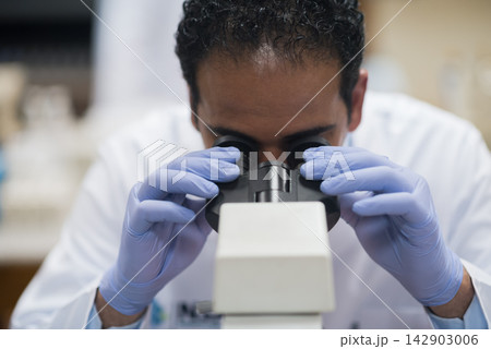 Middle Eastern medical technician looking through microscope in clinical blood testing laboratory wearing gloves and lab coat 142903006