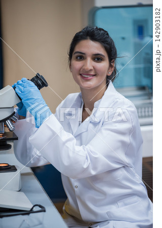 Middle Eastern female scientist in lab coat smiling while using microscope in blood testing laboratory 142903182