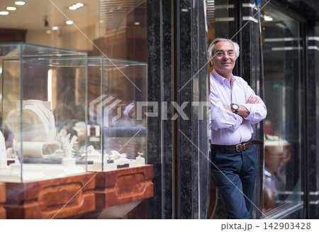 Confident jewelry shop owner standing by glass display as couples, family and friends shop for wedding rings Confident jewelry shop owner standing by glass display as couples, family and friends shop for wedding rings 142903428