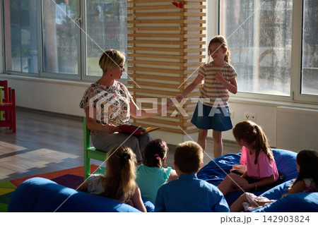 Teacher reading to children in a cozy classroom circle while a girl shares with the group Teacher reading to children in a cozy classroom circle while a girl shares with the group 142903824