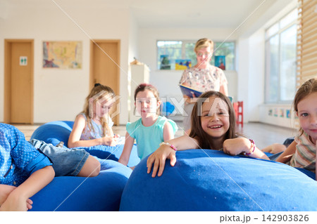 Happy children and teacher reading together on blue beanbags in a bright classroom Happy children and teacher reading together on blue beanbags in a bright classroom 142903826