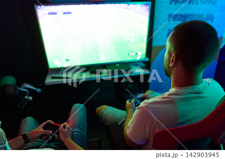Two Friends Playing Console Video Game Together on TV in Dimly Lit Gaming Room 142903945
