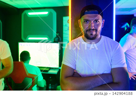 Confident Male Gamer Standing in Neon Esports Gaming Room With Team Playing in Background Confident Male Gamer Standing in Neon Esports Gaming Room With Team Playing in Background 142904188
