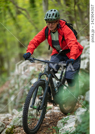 Experienced cyclist mountain biking on a rocky forest trail wearing red jacket and helmet 142904307