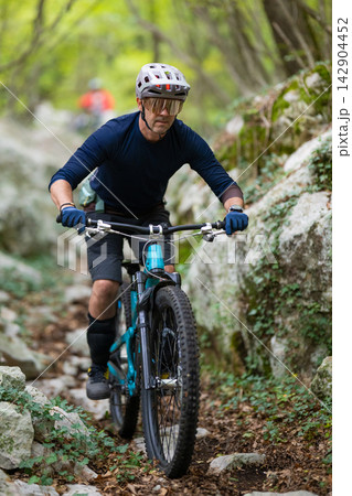 Male mountain biker riding a blue full-suspension bike on a rocky forest trail in summer 142904452