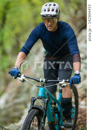 Experienced mountain biker riding a teal full suspension bike on a forest trail wearing helmet and protective gear Experienced mountain biker riding a teal full suspension bike on a forest trail wearing helmet and protective gear 142904453