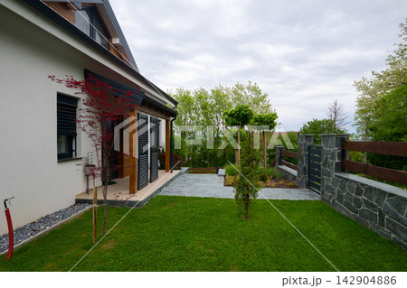 Modern suburban house with landscaped front yard, stone patio and wooden fence under cloudy sky 142904886