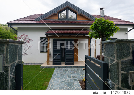 Modern suburban house with gated stone entrance, covered porch and shuttered front door under pitched roof Modern suburban house with gated stone entrance, covered porch and shuttered front door under pitched roof 142904887