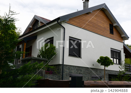 Modern suburban house exterior with wood siding, stone foundation and landscaped yard under overcast sky Modern suburban house exterior with wood siding, stone foundation and landscaped yard under overcast sky 142904888