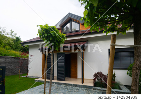 Modern suburban house with covered entrance, wooden shutters and landscaped front yard with trees Modern suburban house with covered entrance, wooden shutters and landscaped front yard with trees 142904939