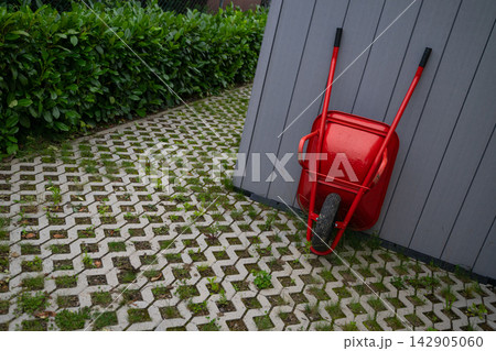 Red wheelbarrow leaning against a modern modular home shed on permeable paver patio with hedge 142905060
