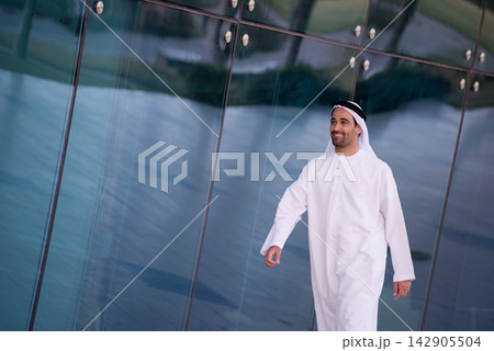 Smiling Arab man in traditional white kandura walking by modern glass building Smiling Arab man in traditional white kandura walking by modern glass building 142905504