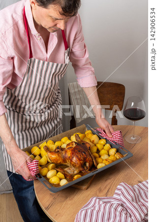 A man prepares a pre-Christmas dinner in his kitchen. He places a tray of baked chicken and potatoes on the table. Surrounding decor hints at a festive atmosphere 142906534