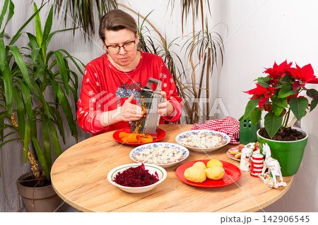 A woman grates carrots in the kitchen for making herring under fur coat salad, the traditional Ukrainian or Russian Christmas salad Shuba. Nearby are bowls with beets, potatoes, and a festive plant A woman grates carrots in the kitchen for making herring under fur coat salad, the traditional Ukrainian or Russian Christmas salad Shuba. Nearby are bowls with beets, potatoes, and a festive plant 142906545