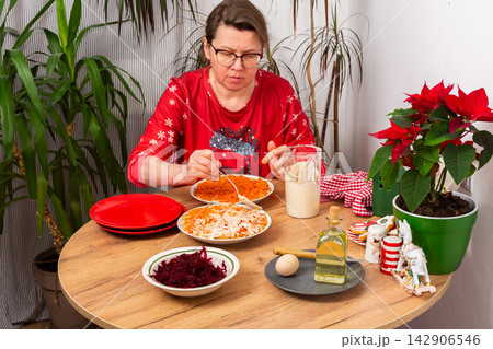 In a cozy home setting, a woman mixes layers of boiled vegetables with mayonnaise to prepare Olivier salad. The scene shows holiday decorations and festive plants nearby In a cozy home setting, a woman mixes layers of boiled vegetables with mayonnaise to prepare Olivier salad. The scene shows holiday decorations and festive plants nearby 142906546