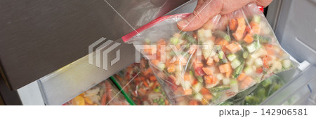A man reaches into the refrigerator and takes out a bag of frozen vegetables. He is preparing to cook during the afternoon in a kitchen setting, banner 142906581