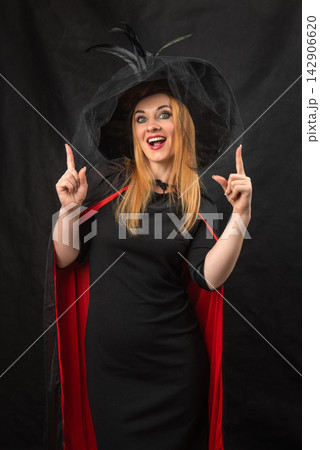 Happy young woman wearing a black witch hat and cape, pointing both index fingers upwards, ready for Halloween festivities on a dark background Happy young woman wearing a black witch hat and cape, pointing both index fingers upwards, ready for Halloween festivities on a dark background 142906620
