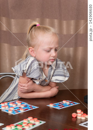 Young blonde girl sits at a wooden table playing a card and token game, concentrating thoughtfully while developing cognitive and strategic skills during quiet indoor playtime Young blonde girl sits at a wooden table playing a card and token game, concentrating thoughtfully while developing cognitive and strategic skills during quiet indoor playtime 142906680