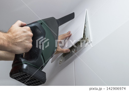 Hands lifting a plastic vent cover and preparing a handheld vacuum for vent cleaning, illustrating home HVAC maintenance task. Hands lifting a plastic vent cover and preparing a handheld vacuum for vent cleaning, illustrating home HVAC maintenance task. 142906974