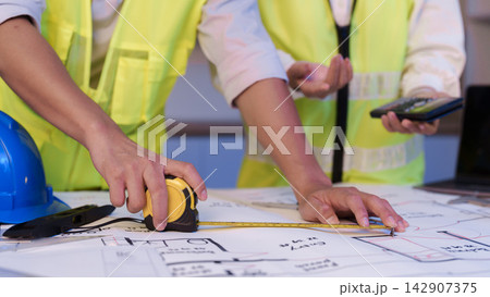 Cropped hands of engineers measuring blueprint with tape measure for construction project 142907375