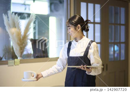 カフェのテーブルにコーヒーを運ぶ女性スタッフ カフェのテーブルにコーヒーを運ぶ女性スタッフ 142907720