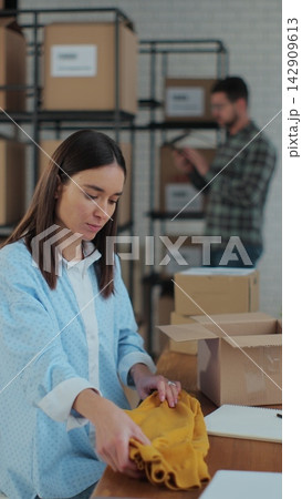 Vertical video. An employee packs a yellow jumper in a room with shelves full of parcels. The woman owner of an online store works on a computer at his desk in a warehouse. Vertical video. An employee packs a yellow jumper in a room with shelves full of parcels. The woman owner of an online store works on a computer at his desk in a warehouse. 142909613