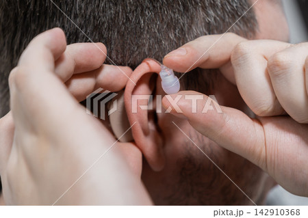 A man places a modern beige hearing aid into his ear, demonstrating careful adjustment of a discreet medical device to improve auditory clarity and support daily communication needs A man places a modern beige hearing aid into his ear, demonstrating careful adjustment of a discreet medical device to improve auditory clarity and support daily communication needs 142910368