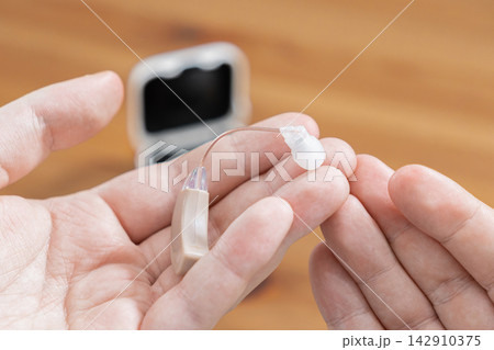 A person holds a small beige hearing aid in an open palm to demonstrate assistive medical technology that enhances daily communication and auditory health for those with hearing loss A person holds a small beige hearing aid in an open palm to demonstrate assistive medical technology that enhances daily communication and auditory health for those with hearing loss 142910375