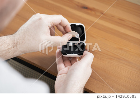 A man holds a hearing aid above a protective case on a wooden desk, emphasizing modern healthcare accessibility and the daily routine of managing auditory support for better wellness A man holds a hearing aid above a protective case on a wooden desk, emphasizing modern healthcare accessibility and the daily routine of managing auditory support for better wellness 142910385
