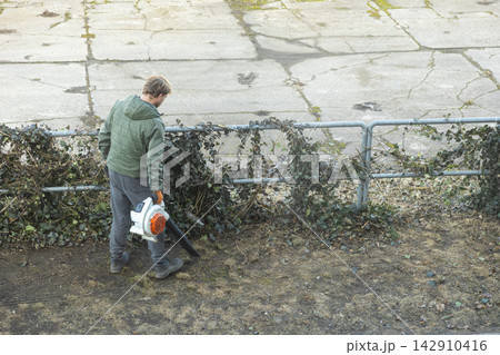 A man uses a power blower to clear dry debris near an ivy covered metal fence, reflecting diligent outdoor maintenance work and professional landscape care in a calm public area 142910416