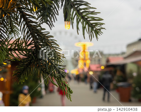 Evergreen branch with warm glowing light bulbs overlooking a festive holiday market featuring a blurred ferris wheel, creating a cozy winter season atmosphere in a European city Evergreen branch with warm glowing light bulbs overlooking a festive holiday market featuring a blurred ferris wheel, creating a cozy winter season atmosphere in a European city 142910438