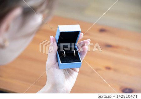 A woman holds an open blue jewelry gift box with sparkling diamond hoop earrings inside, capturing an intimate moment of surprise and elegance in a warm wooden interior home setting A woman holds an open blue jewelry gift box with sparkling diamond hoop earrings inside, capturing an intimate moment of surprise and elegance in a warm wooden interior home setting 142910471