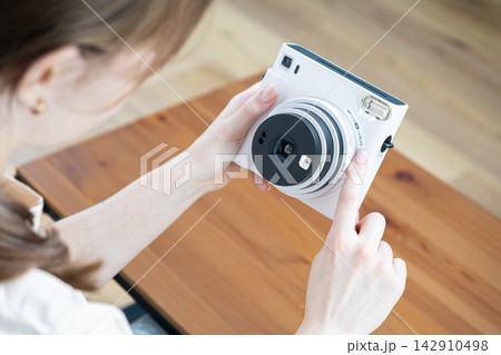 A young woman adjusts the lens on a white instant camera while sitting at a wooden table, capturing a casual and creative moment during a lifestyle hobby session at home indoors today A young woman adjusts the lens on a white instant camera while sitting at a wooden table, capturing a casual and creative moment during a lifestyle hobby session at home indoors today 142910498