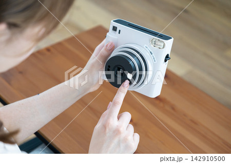 A young person adjusts a white instant camera while sitting at a rustic wooden table, capturing a creative hobby moment with a modern device designed for vintage style photography A young person adjusts a white instant camera while sitting at a rustic wooden table, capturing a creative hobby moment with a modern device designed for vintage style photography 142910500