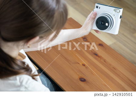 A young person holds a white instant camera to take a selfie over a wooden desk, capturing a creative moment of self expression with modern analog technology in a bright home A young person holds a white instant camera to take a selfie over a wooden desk, capturing a creative moment of self expression with modern analog technology in a bright home 142910501