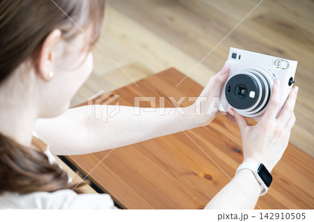 A young woman holds a white instant camera to capture a photograph at home, evoking nostalgic hobbies, creative lifestyle, and the joy of preserving memories through analogue tech 142910505